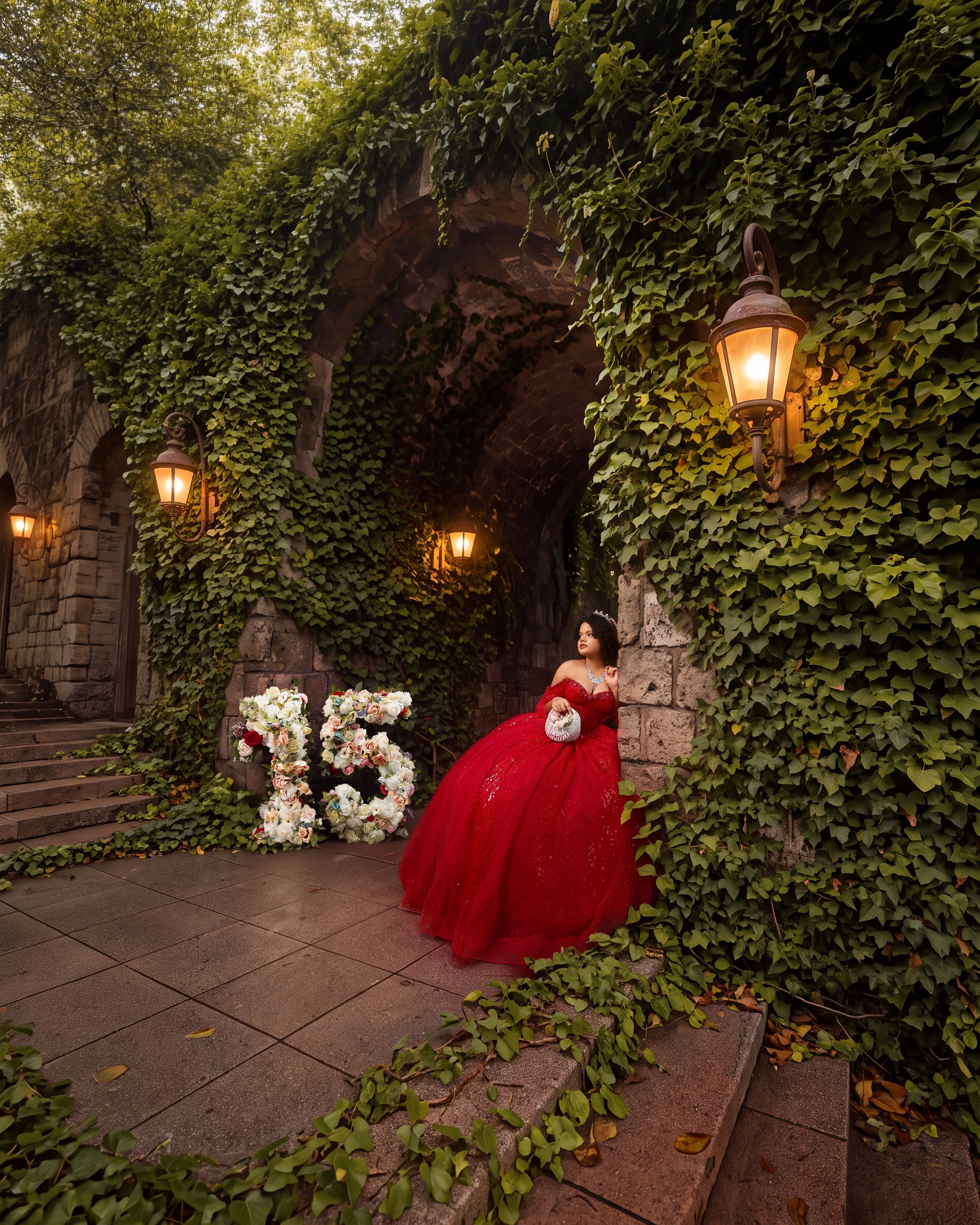 Quinceañera con vestido rojo bajo un arco de piedra con hiedra y faroles, junto a un número 15 de flores