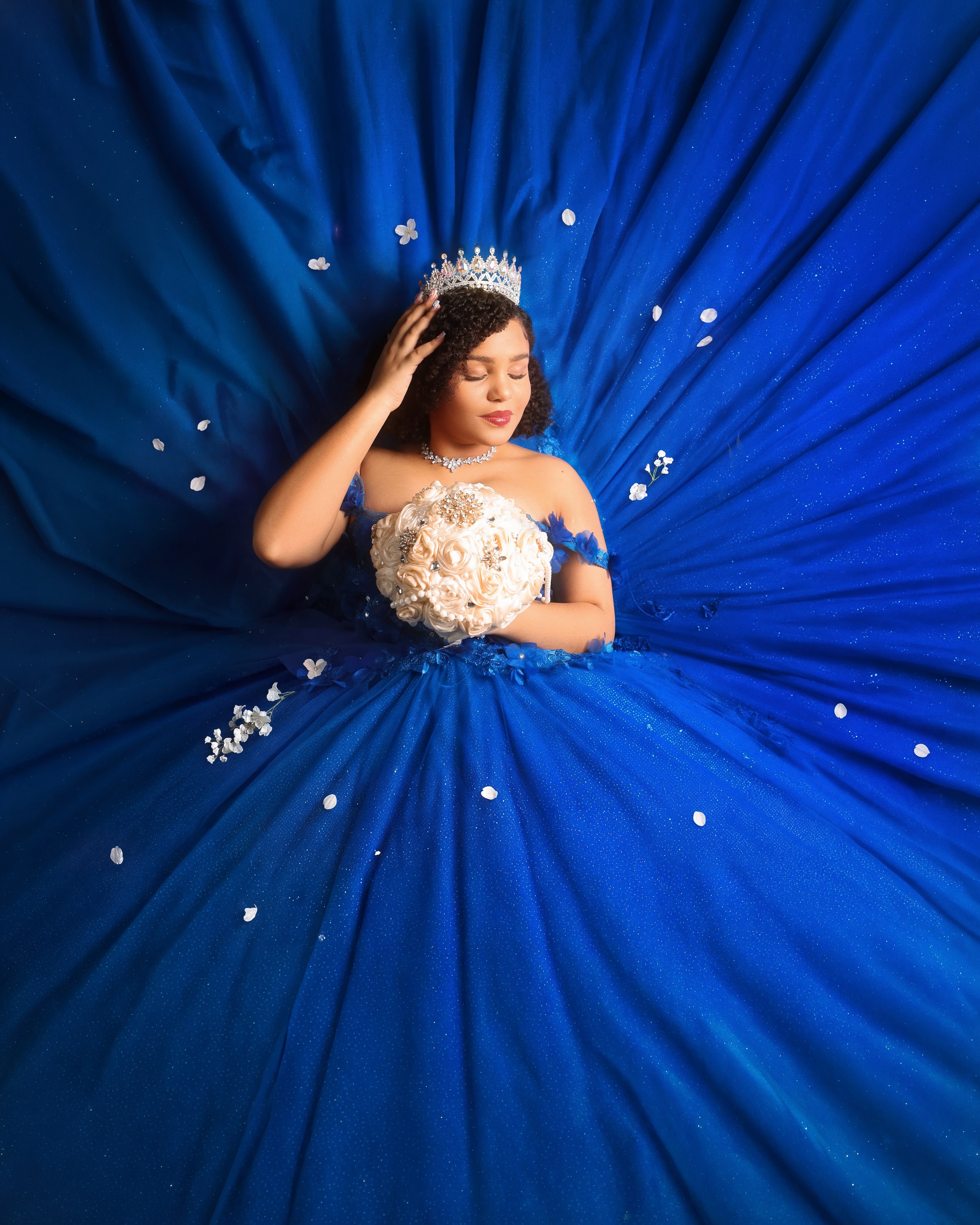 Quinceañera con corona recostada sobre su vestido azul real extendido, vista desde arriba
