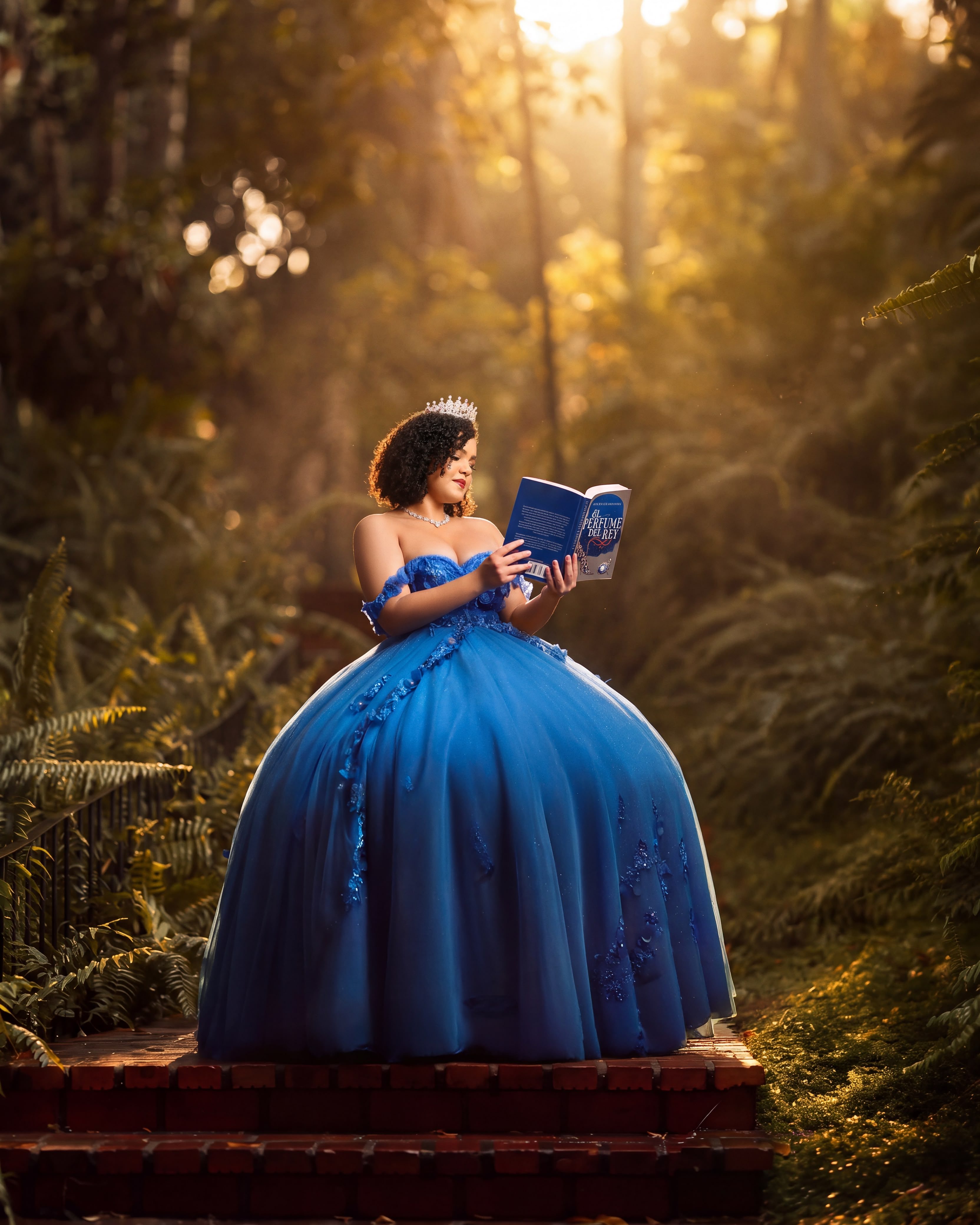 Quinceañera con vestido azul real leyendo un libro en un bosque iluminado por el sol