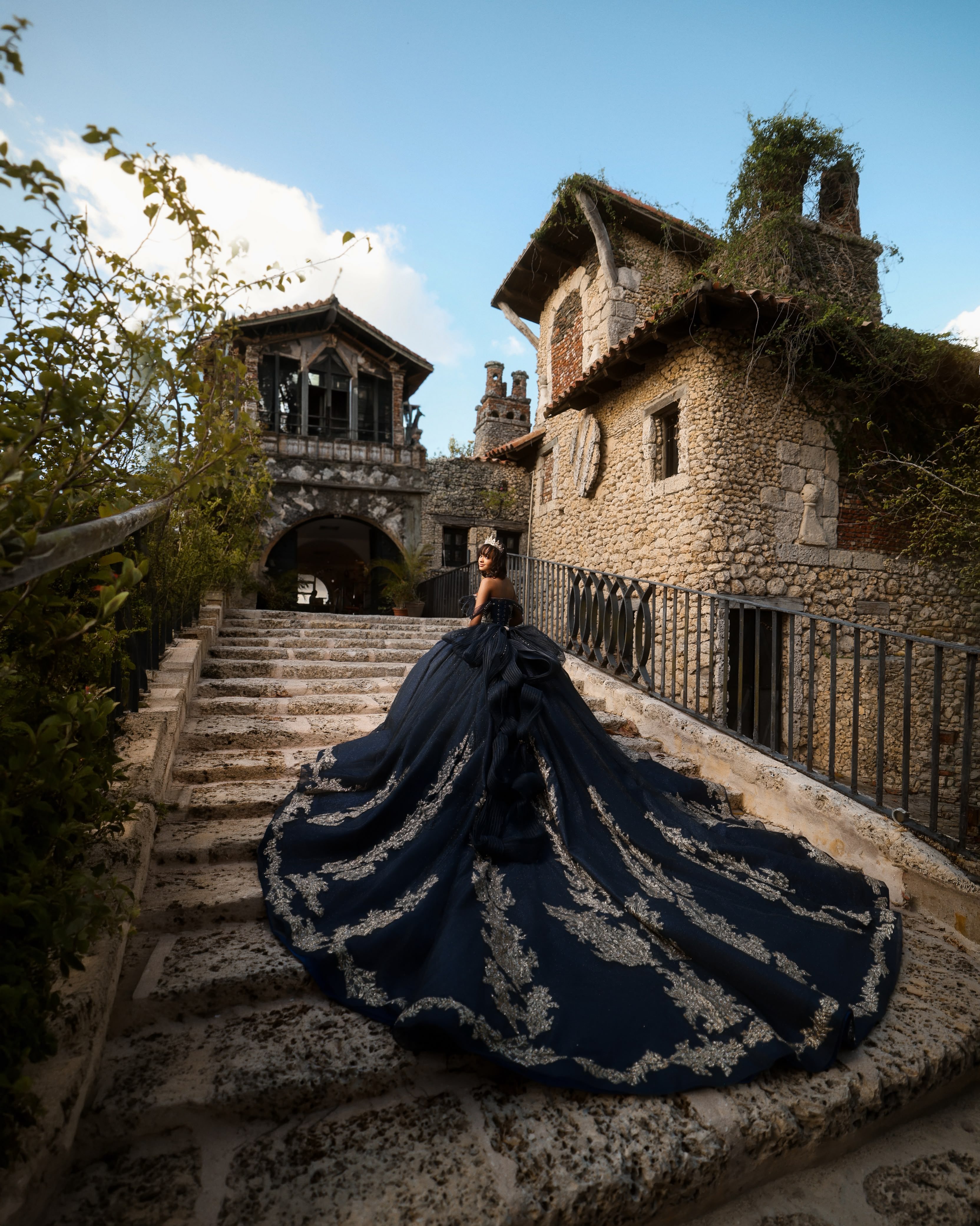 Quinceañera con vestido azul marino de cola bordada en una escalinata de piedra junto a un castillo