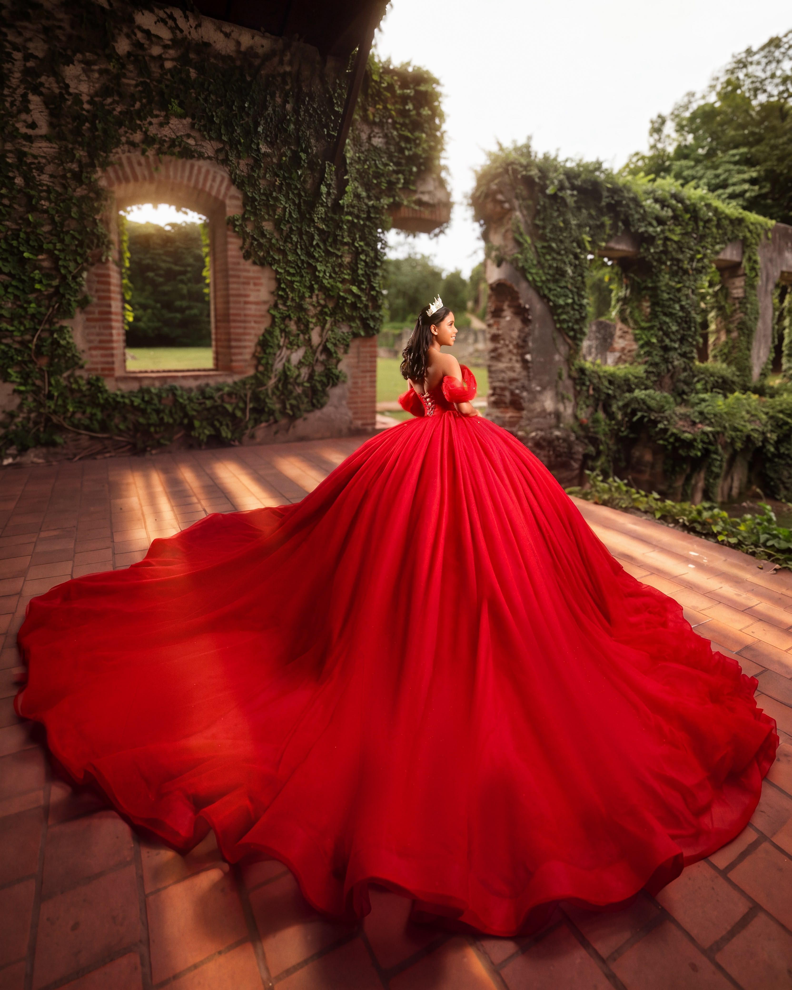 Quinceañera de espaldas con vestido rojo de larga cola en unas ruinas cubiertas de hiedra