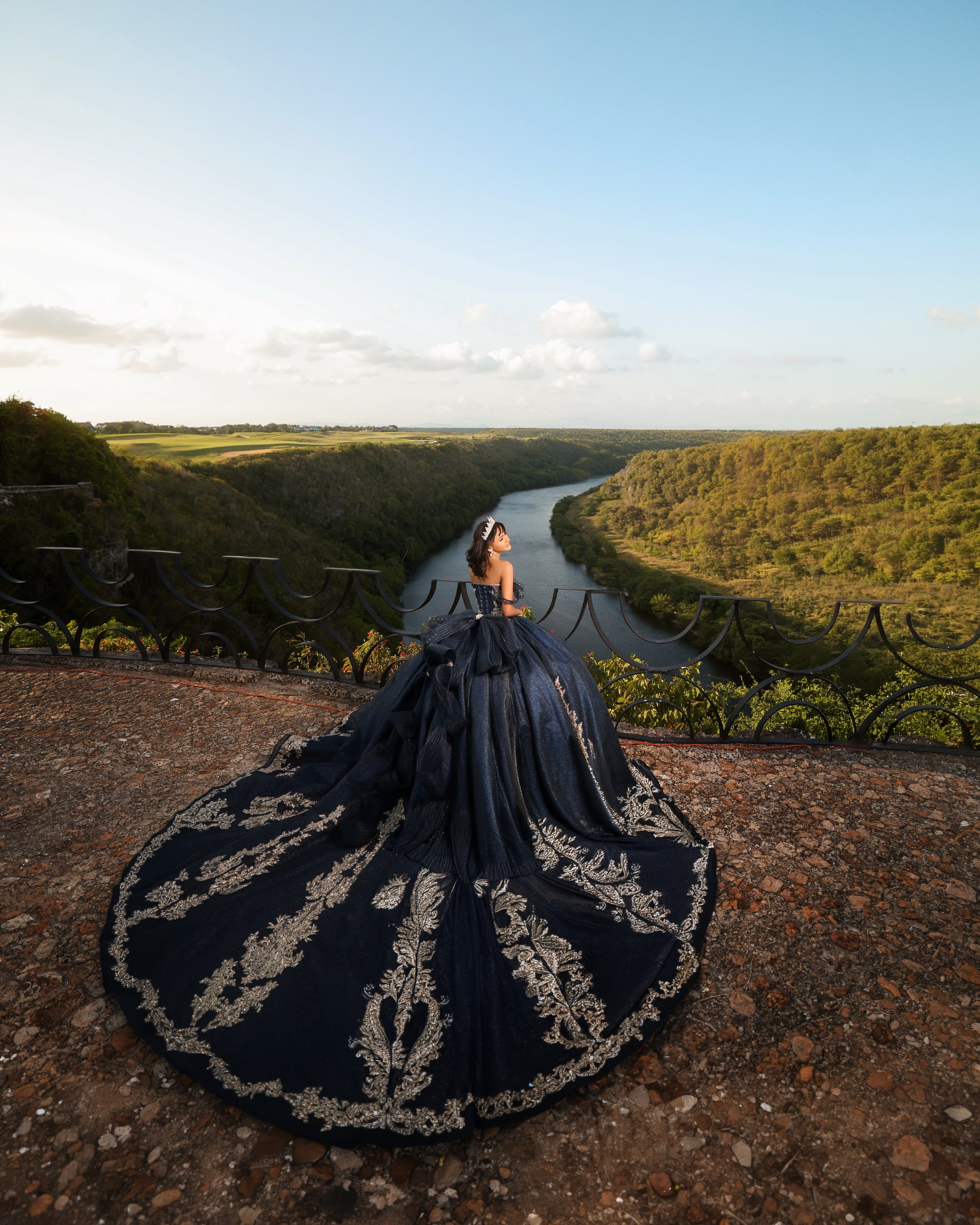 Quinceañera de espaldas con vestido azul marino bordado en un mirador sobre un río y un cañón verde