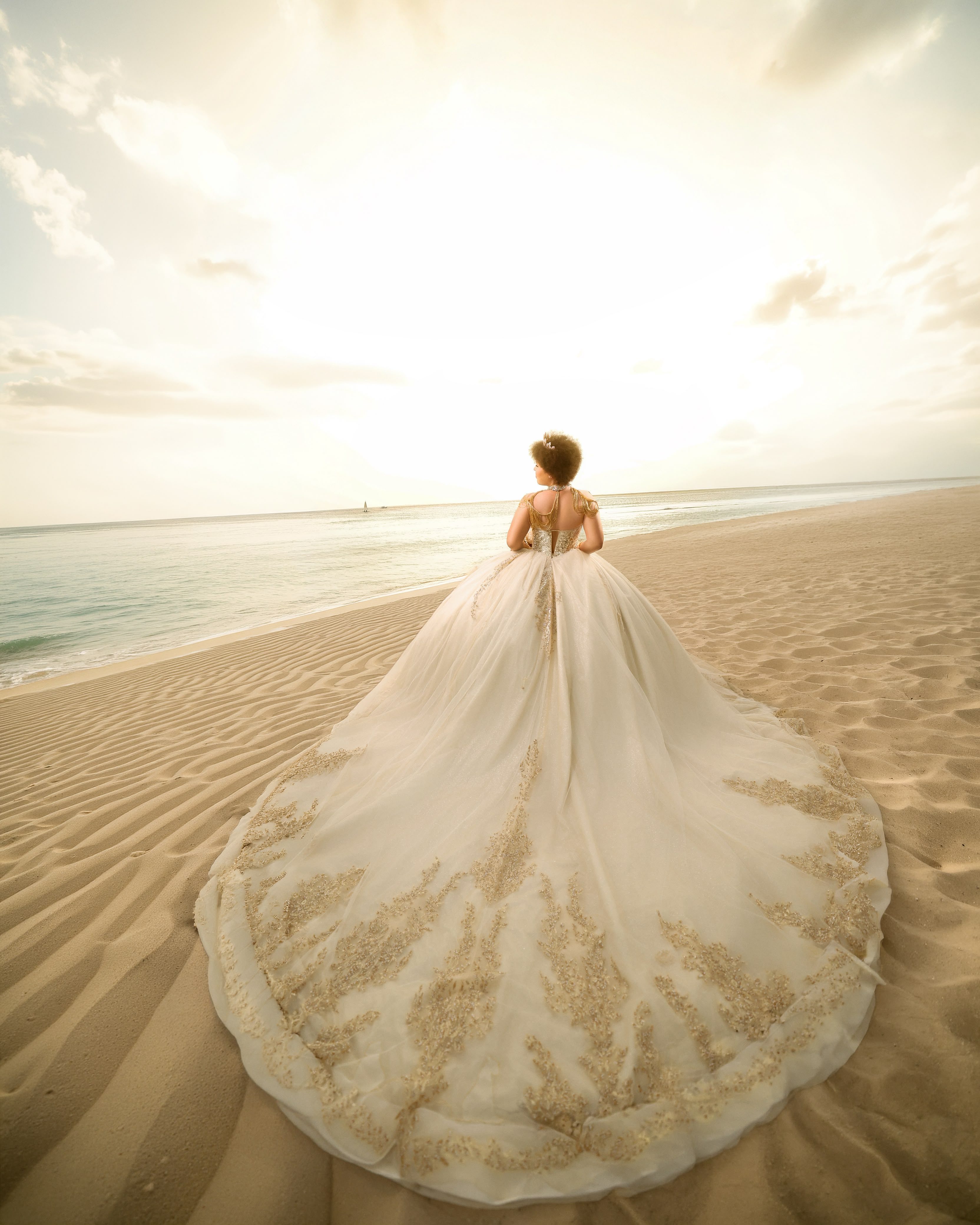 Quinceañera de espaldas con vestido marfil de cola bordada en dorado sobre la arena de la playa