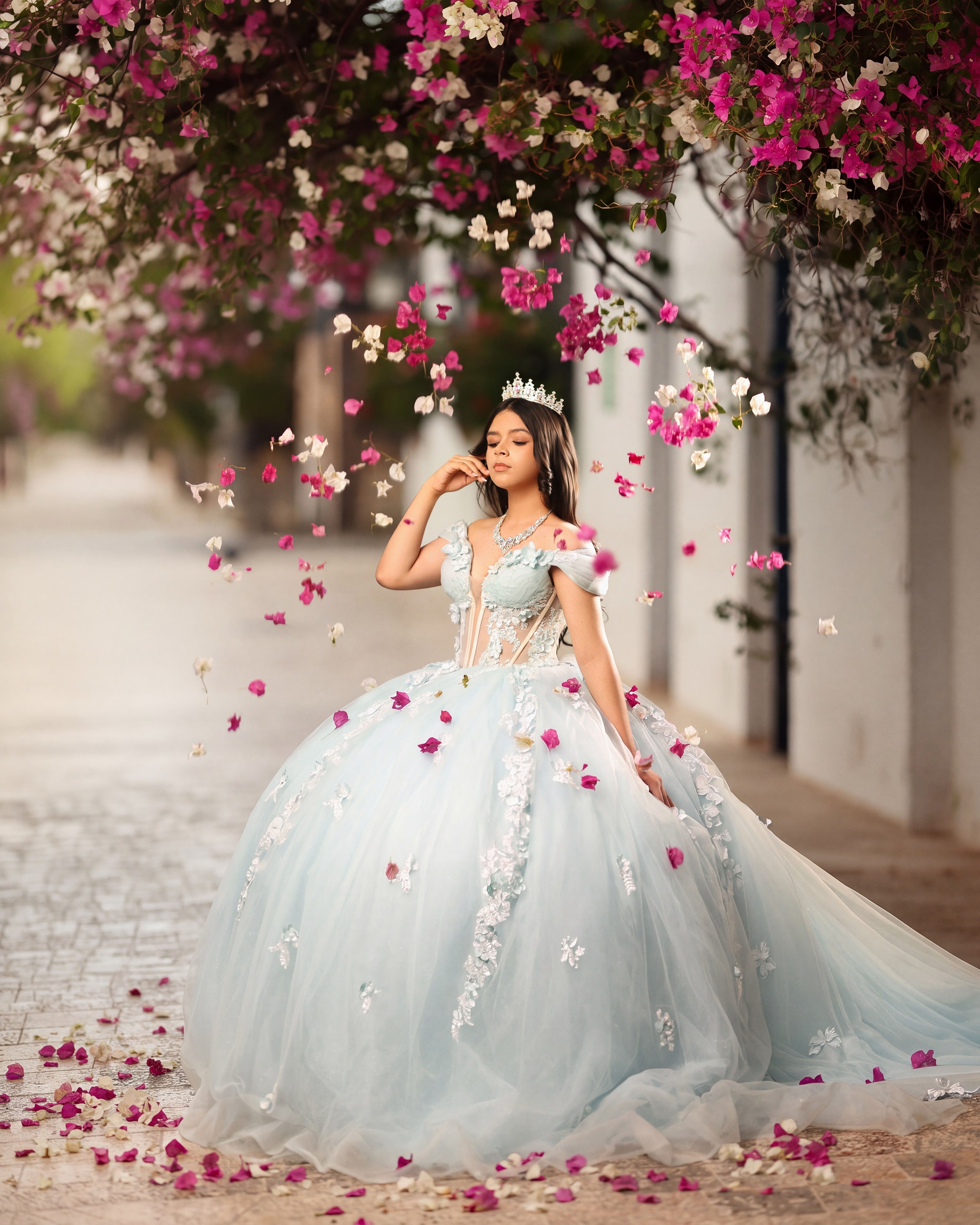 Quinceañera con vestido celeste bajo una lluvia de pétalos de buganvilia en una calle colonial