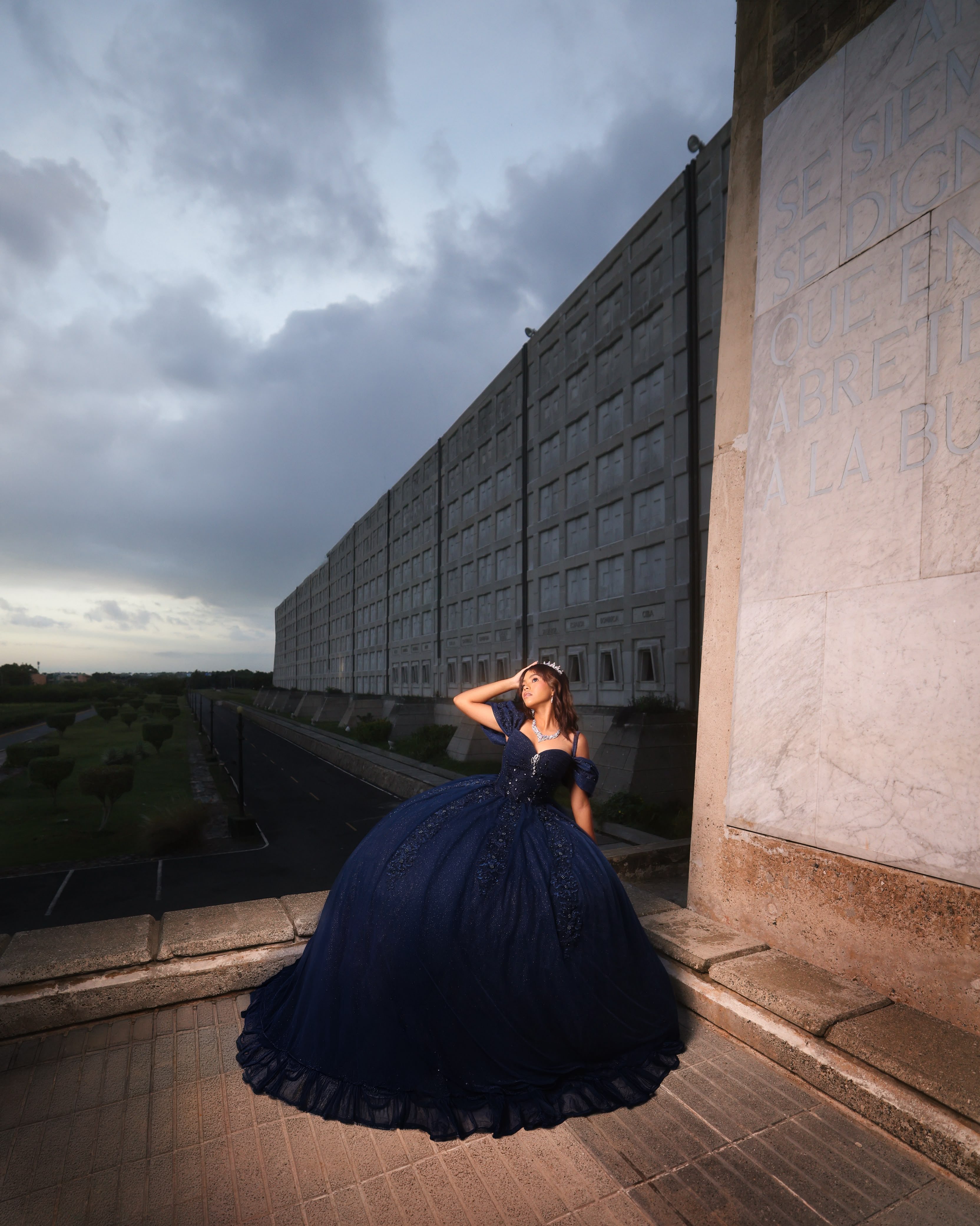 Quinceañera con vestido azul marino junto a un muro monumental bajo un cielo de tormenta