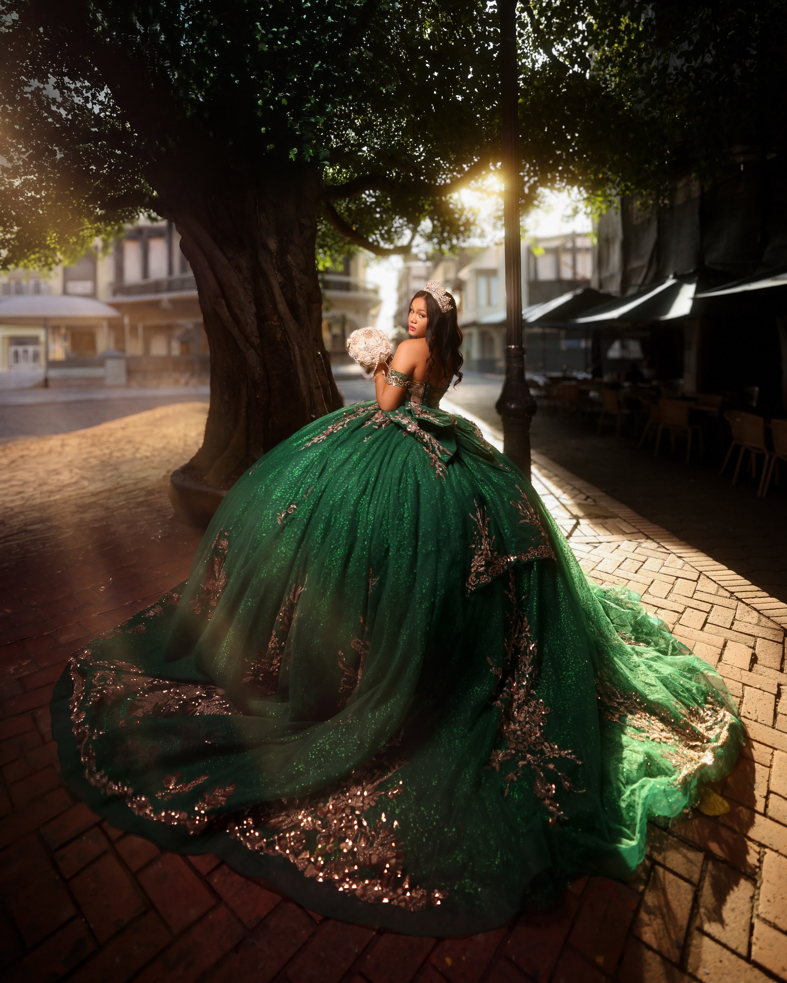 Quinceañera con vestido verde esmeralda bordado bajo un árbol en una calle colonial