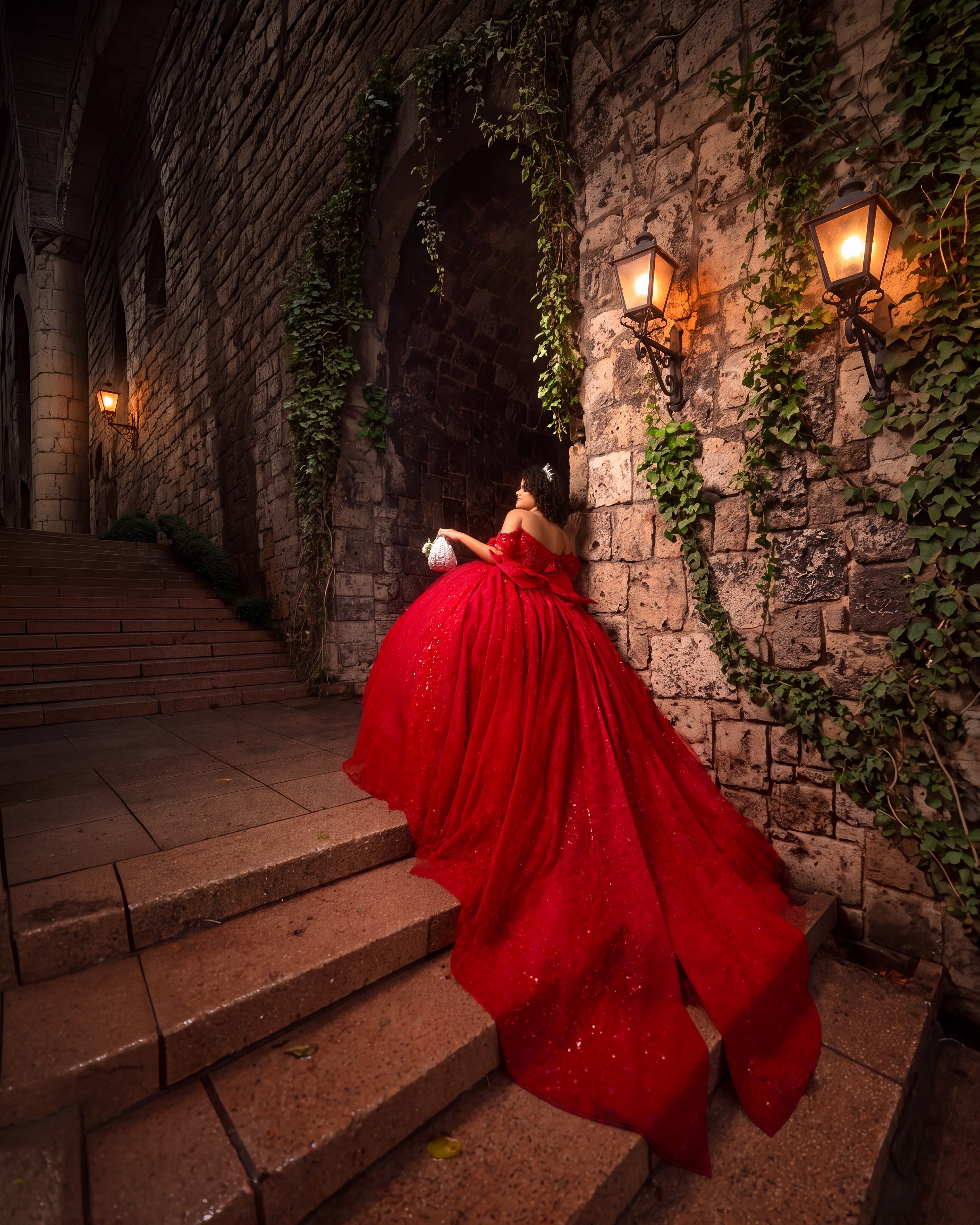 Quinceañera con vestido rojo de cola en una escalera de piedra con faroles encendidos