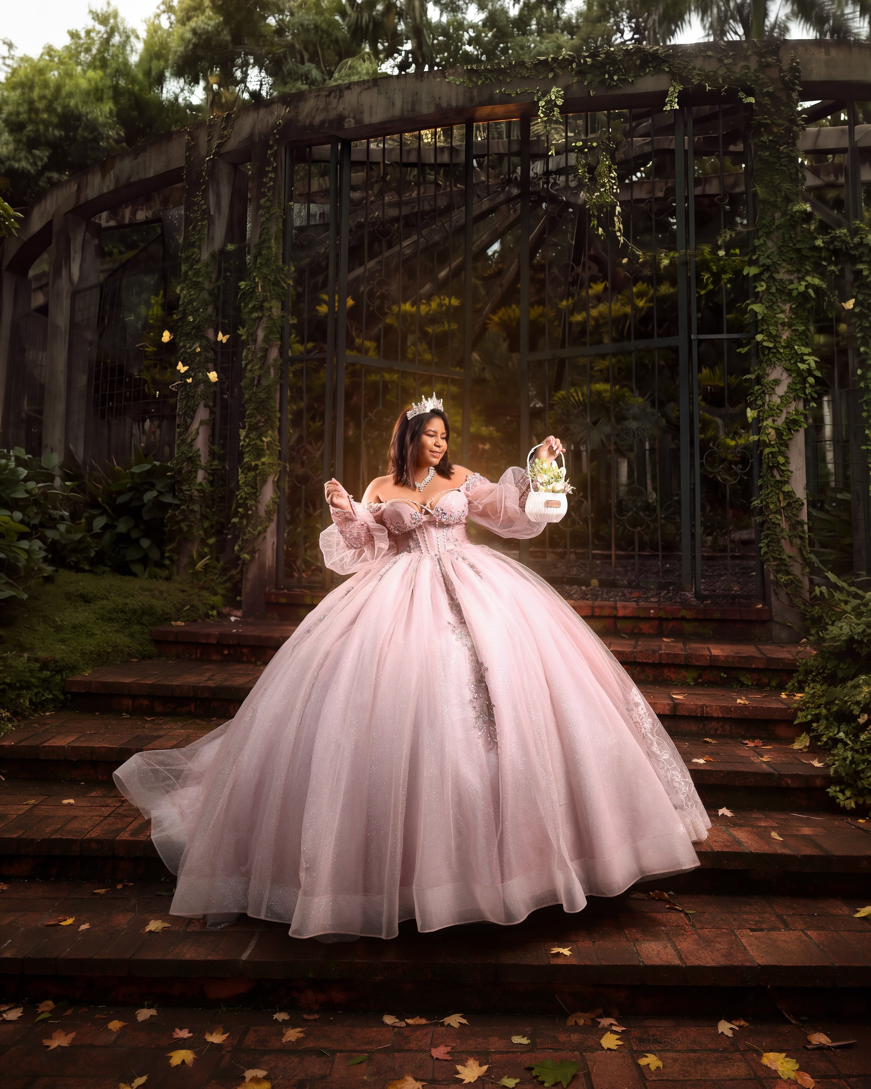 Quinceañera con vestido rosa de mangas abullonadas y una canasta de flores frente a una pérgola de hierro